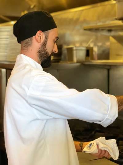 Chris Mills, wearing chefs whites and a black chef's beanie, pours oil into a pan over the gas range in the Davio's kitchen