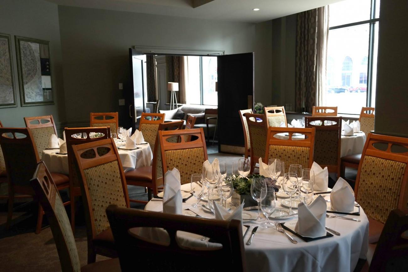 Light shines through tall windows on a room with multiple round tables set for 6 each and decorated with white tablecloths and greenery in centerpieces. The couches in the lounge area of the main dining room are visible through the open doors.